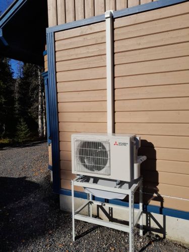 Ilmalämpöpumpun ulkoyksikkö jalustalle asennettuna puuvuorauksiseen talon seinään.